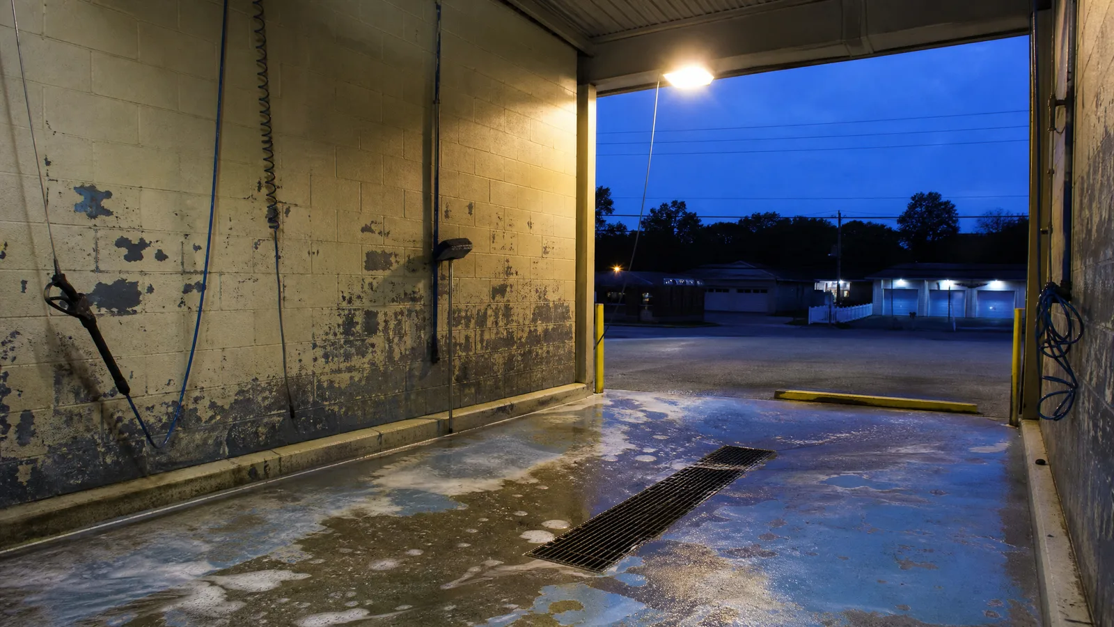 Empty self-service car wash bay at dusk, foam on the wet floor, wand and coiled hose on the block wall